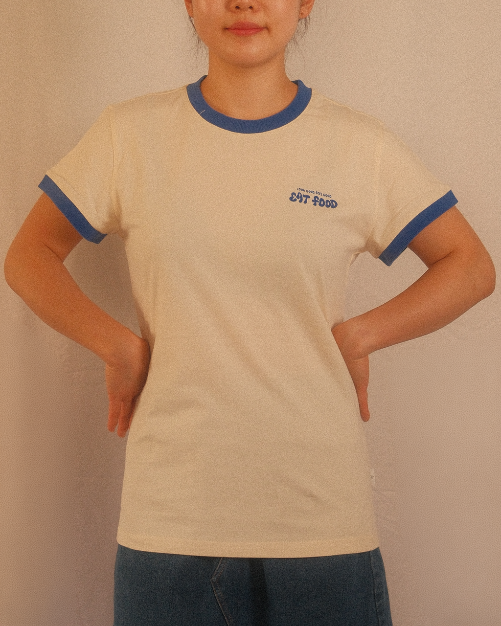 Person wearing a beige regular fit t-shirt with blue trim and text, standing against a plain background