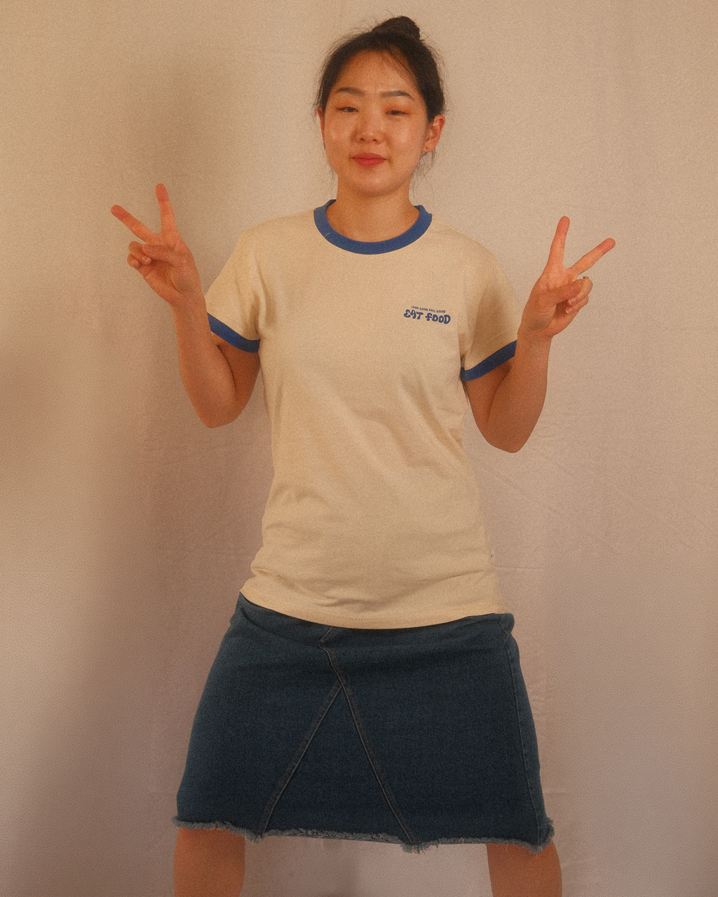 Person wearing a beige regular fit t-shirt with blue trim and a denim skirt, making peace signs against a plain background.